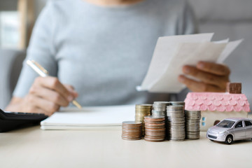 Close up of hand holding bills while writing, stack of coins, toy house and car on table, saving for future, manage to success, business and finance concept.