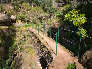 A steel cable safety fence protects tourists and hikers from a steep drop on a lavada walking trail near Monte in Funchal district of Madeira. Taken on a sunny day.