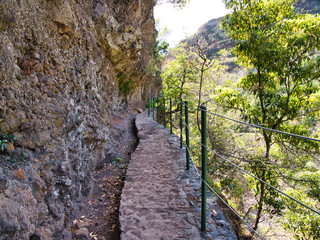 A steel cable safety fence protects tourists and hikers from a steep drop on a lavada walking trail near Monte in Funchal district of Madeira. Taken on a sunny day.