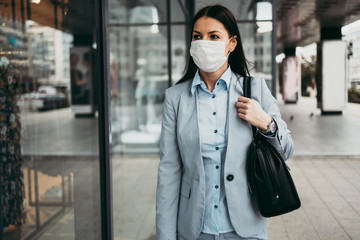 Young and elegant business woman with face protective mask standing on empty street and looking at...