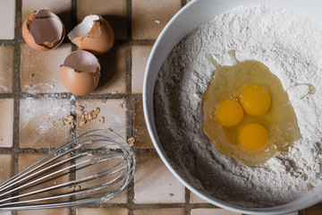 A mixing bowl is filled with plain white flour and three eggs as the eggshells are still next to the bowl and a whisk can be seen in the bottom left corner
