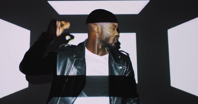 African American attractive man in black leather jacket dancing and posing to camera on white wall background with graphic squares projections. Projected figures moving on handsome male model dancer.