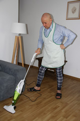 55 year old bald man in home clothes and apron vacuuming floors in a room near the sofa, concept of distribution of household chores in the family