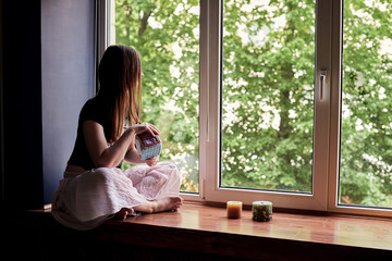 Obraz na płótnie Canvas Young brunette woman, wearing black top and light pale pink yoga pants, sitting on windowsill with colorful aroma candles, looking through at green trees in park, thinking, meditating.