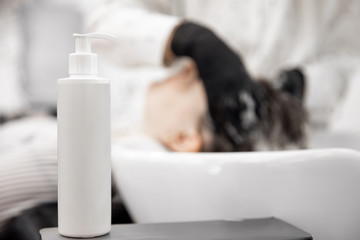 Close-up white plastic bottle for shampoo, background woman washes head in beauty salon. Concept...