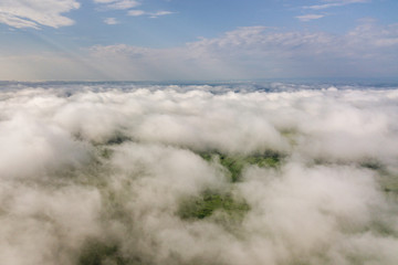 Fototapeta premium Aerial view of white clouds above a town or village with rows of buildings and curvy streets between green fields in summer. Countryside landscape from above.