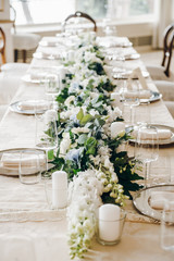 White and green flower composition, candles, plates with napkins, there are on a wedding table covered with tablecloth