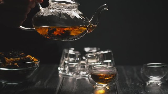 Tea ceremony. The woman's hand pours hot black tea with flowers from a transparent teapot into glass piala cups. Black background, counter light.