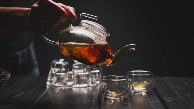Tea ceremony. The woman's hand pours hot black tea with flowers from a transparent teapot into glass piala cups. Black background, counter light.