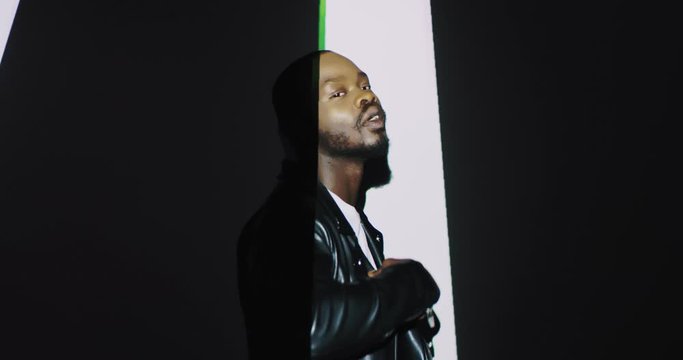 Portrait of stylish young African American male model in black leather jacket dancing on white wall background. Projection of lights, white and black stripes and colours on man. Graphic moving.