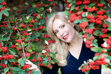 A waist up portrait of Caucasian woman with long blond hair with red near a bush with red berries