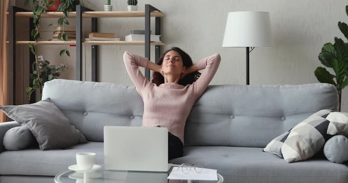 Happy young woman freelancer takes break relaxing from distance job, remote computer work sits on sofa at home office. Smiling lady stretching neck, holding hands behind head enjoys no stress concept.