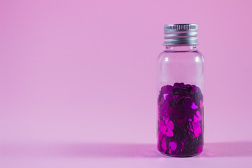 Glass bottle with purple heart shaped sparkles on pink background with copy space. Festive concept for your creative design - obrazy, fototapety, plakaty