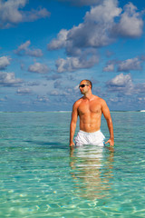 Summer Travel Vacation. Handsome Man With Sexy Body In Fashionable Sunglasses Sunbathing, Tanning At Sea Beach. Fitness Male Model With Sunscreen Lotion, Sun Block Skin Protection Cream In Pocket