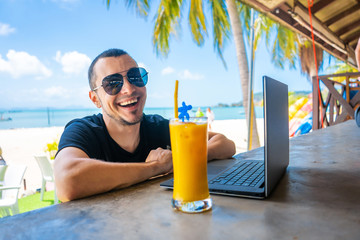 The guy works at a laptop, sitting in a beach bar and drinks a mango shake. Successful business. Remotely work.