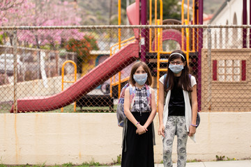 Two school aged girls are in front of the closed and locked school during the covid-19 outbreak. They are worried and disappointed while wearing face masks. 