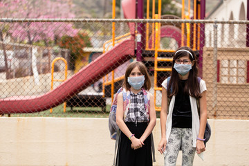 Two school aged girls are in front of the closed and locked school during the covid-19 outbreak. They are worried and disappointed while wearing face masks. 