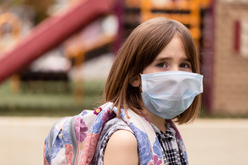 A school aged girl is longing to attend her closed school. She is in front of her school with a facemask on during the covid-19 outbreak.