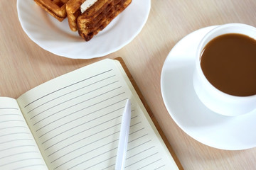 Top view: two pieces of fresh waffles with cream on a plate with morning strong coffee and an open notebook on the table