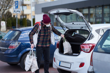 Caucasian man wearing medical mask packs huge amount of bags with food into car after shopping...