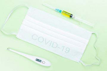COVID-19 concept picture with medical face masks, digital thermometer with high temperature, pills and syringe on the light green background.