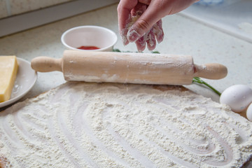 the baker sprinkles flour on the table. Eggs, rolling pin, bowl with sauce and cheese.