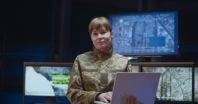 Portrait shot of Caucasian beautiful young woman from army standing in monitoring room and using laptop computer. Female soldier looking straight and smiling to camera.