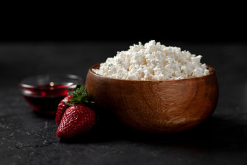 tasty homemade cottage cheese in a wooden plate with strawberries and jam on a dark background, healthy breakfast on a black table