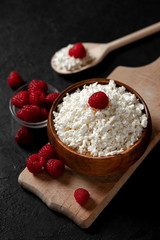 fresh homemade cottage cheese in a wooden bowl with raspberries on a dark background, healthy food on a black table