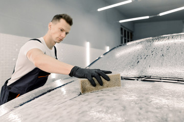 Car washing concept. Young concentrated Caucasian male worker holding the sponge in hand and...