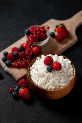 homemade fresh cottage cheese with strawberries, raspberries, blueberries, currants in a wooden plate against a dark background, healthy milk food with wild berries on a black table