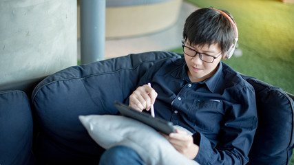 Asian businessman with headphones listening to music working with digital tablet on comfortable sofa at home office. Man with glasses doing online conference on mobile app. Work from home concept