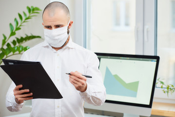 A man in a medical face mask against the coronavirus (COVID-19). An office worker holding a pen and a folder at his workspace with computers and green plants in the background. Coronavirus quarantine