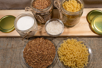 On the board are cans of buckwheat, pasta and sugar. In the bowl lies rice. Buckwheat and pasta also lie on plates.