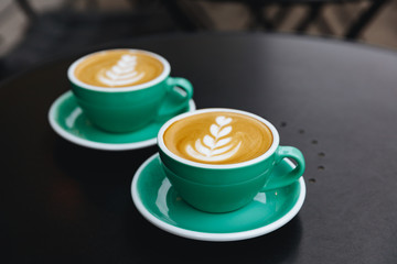 Black table of coffee shop terrace with couple of turquoise colored mugs of freshly brewed cappuccino with lush milk foam. Concept of latte art and barista art
