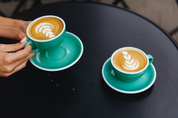 Close up of woman hands with trendy manicure holding one green cup of tasty aroma cappuccino with lush milk foam while another cup is standing near on black table.