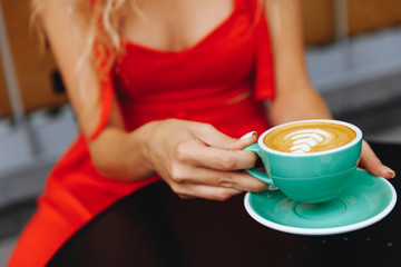 Close up of young girl with blond curly hair wearing trendy red summer dress sitting at black table outdoors and holding turquoise colored cup of aroma cappuccino with latte art in a from of leaf.