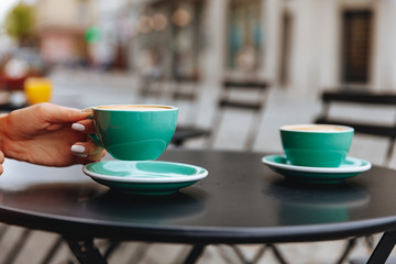 Black metal table with couple of trendy blue colored cups filled with hot freshly brewed cappuccino. Young lady sitting on coffee shop terrace  and going to drink aroma coffee