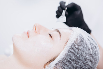Naklejka premium Hands of a Caucasian cosmetologist woman applying a facial mask on a beautiful young Caucasian woman's face lying on the massaging bed