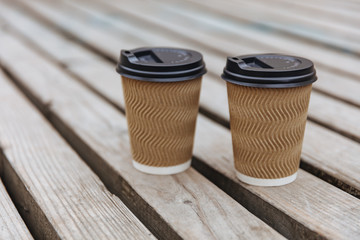 Two disposable paper cups of coffee to takeaway on wooden floor outside the cafe. Hot beverage in mugs with black lids. Morning breakfast on fresh air