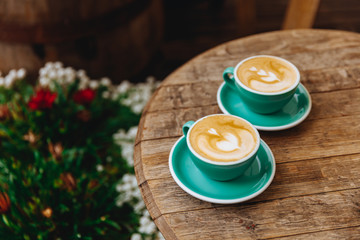 Light blue cups of hot fresh coffee with latte art in form of heart  on wooden table with blur background of flower bush. Retro, vintage effect and copy space