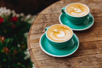 Freshly brewed coffee with heart shape on top in two turquoise colored mugs on saucers places on wooden round table. Hot morning drinks served for couple in love