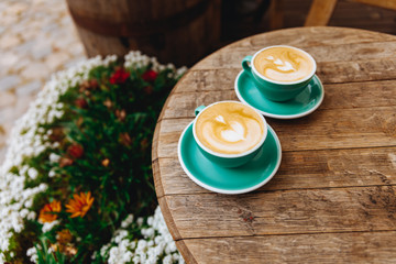Two greenery ceramic cups on saucers filled with hot aroma cappuccino with latte art in a form of heart served on wooden table of summer terrace. Concept of barista art and morning beverage