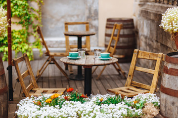 Fototapeta na wymiar Comfortable summer terrace decorated with flowers and wooden barrels where lovely couple can drink hot aroma cappuccino made by professional barista. Two greenery cups of coffee and glasses of water.