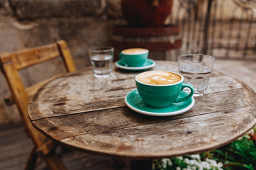 Wooden round table of cafe terrace with two greenery ceramic cups of  hot cappuccino with latte art made by barista and served with glass of water. A cozy place for coffee break in noisy city