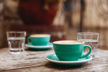 Tasty aroma cappuccino with latte art on top in light blue ceramic cups with glasses of water over blur background. Concept of breakfast and morning beverage