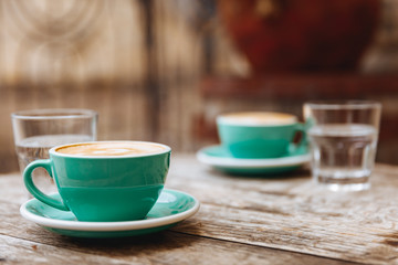 Obraz na płótnie Canvas Two turquoise colored cups of freshly brewed aroma coffee with lush milk served with glasses of water on round wooden table at cafe. Vintage effect and cozy atmosphere