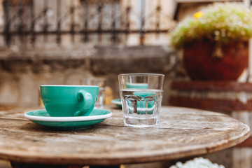 Greenery ceramic cup of cappuccino coffee on wooden round table with glass of water at summer terrace. Enjoyment of hot aroma beverage on fresh air.