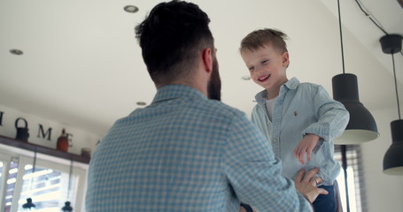 Low angle happy cute little son enjoying fun jumping into young father's arms from house kitchen...