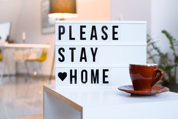 Stay home concept. Self quarantine during Covid-19 coronavirus pandemic. PLEASE STAY HOME written on lightbox in bright home interior with bokeh.